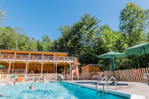 A sunny pool scene with people swimming and a person jumping in, surrounded by trees and a wooden building.
