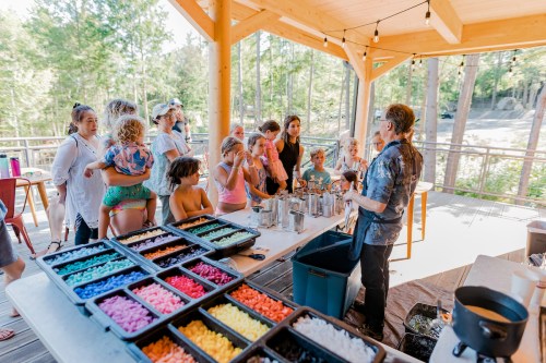 A group of children and adults gather around a table with colorful materials, engaging in a creative activity outdoors.