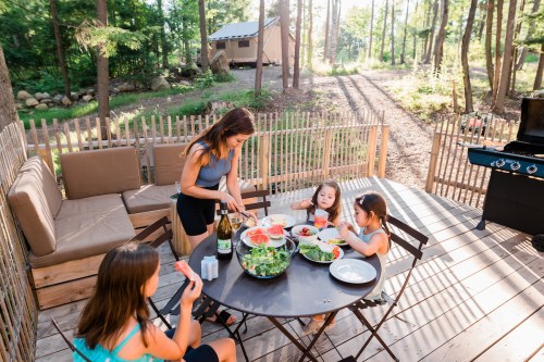 A woman prepares food at a picnic table while three girls enjoy a meal outdoors in a forested area.