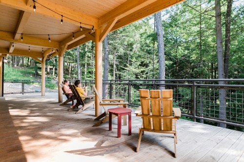 Two people relax on a wooden porch surrounded by trees, with chairs and a railing overlooking a serene forest view.