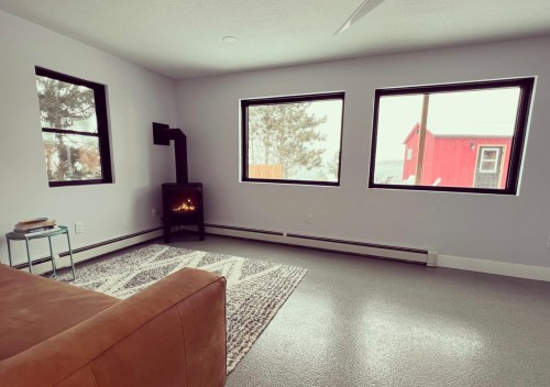 Cozy living room with a brown couch, a small stove, and large windows showing a snowy landscape outside.