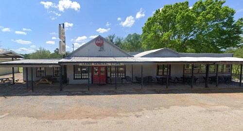 A rustic general store with a red door, surrounded by trees and blue skies, featuring outdoor seating.