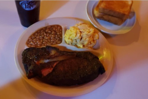 A plate with a large steak, baked beans, scrambled eggs, and toast, alongside a glass of Dr Pepper.