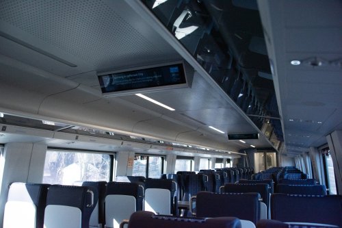 Interior of a train carriage with empty seats and digital display screens above. Bright, modern design.