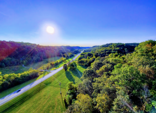 A scenic view of a winding road through lush green hills under a bright blue sky and sun.