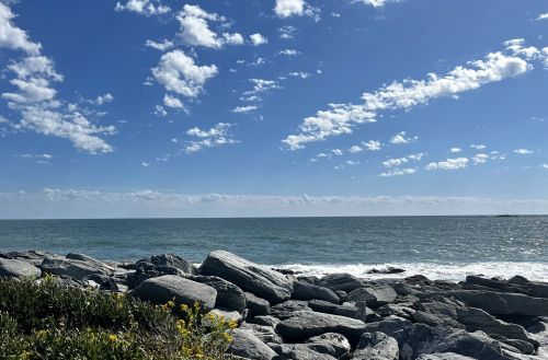 A rocky shoreline meets a calm sea under a bright blue sky with scattered clouds.