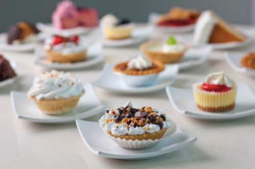 A variety of colorful desserts on white plates, including mini pies, cupcakes, and cream-topped treats.