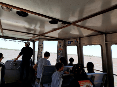 A group of people inside a boat, with a view of the river through large windows. A person is steering at the front.