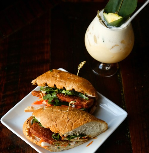 Two crispy sandwiches filled with greens and vegetables, served on a white plate alongside a tropical drink.