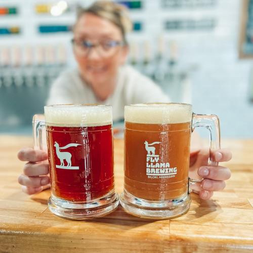 A person holds two mugs of craft beer in a brewery, showcasing a red and a brown beer with a logo on the glass.