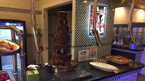 A chocolate fountain with cascading chocolate, surrounded by snacks and a sign about food waste.