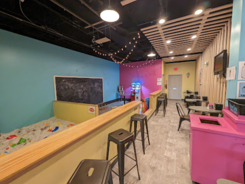 Brightly colored play area with a ball pit, seating, and arcade games, featuring a chalkboard and playful decor.