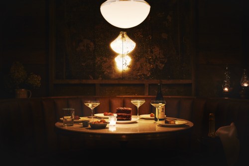 A dimly lit table set with cocktails, dessert, and snacks, illuminated by a warm pendant light.