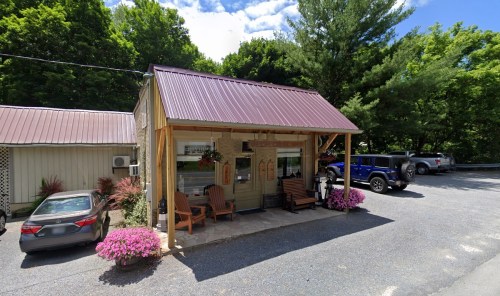 A small building with a metal roof, surrounded by greenery and flowers, featuring outdoor seating and parked cars.