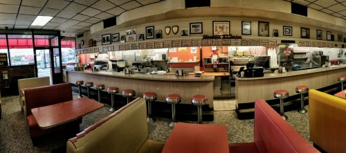 A retro diner interior with a long counter, red stools, and colorful booths, featuring vintage photos on the walls.