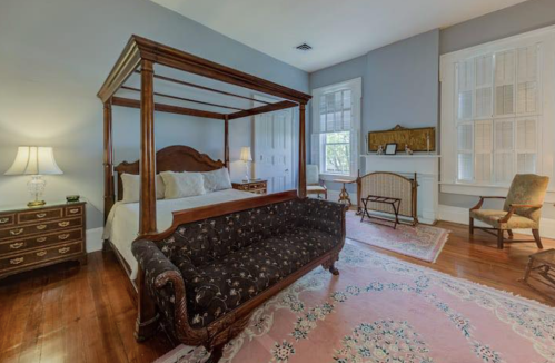 A spacious bedroom featuring a four-poster bed, vintage furniture, and a patterned area rug. Natural light from windows.