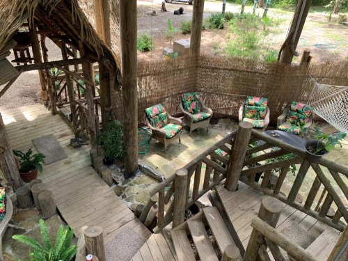 A cozy outdoor seating area with floral chairs, wooden stairs, and a hammock surrounded by greenery.