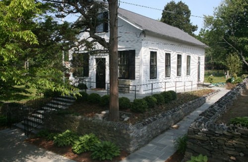 A white, historic house with a gray roof, surrounded by greenery and a stone pathway, set in a sunny outdoor area.