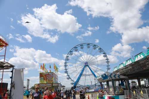 A vibrant fair scene with a large Ferris wheel, colorful stalls, and people enjoying a sunny day under fluffy clouds.