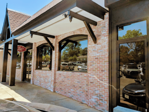 Exterior view of a brick building with large windows and a sign, surrounded by parked cars and trees.