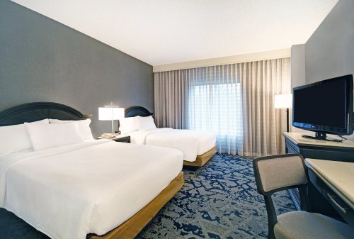 A modern hotel room with two beds, a desk, and a large window with sheer curtains.