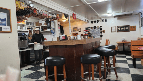 A cozy diner interior with a wooden bar, stools, and a staff member in the background. Black and white checkered floor.