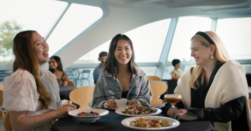 Three women enjoy a meal together at a stylish restaurant, laughing and sharing drinks, with a bright, modern backdrop.