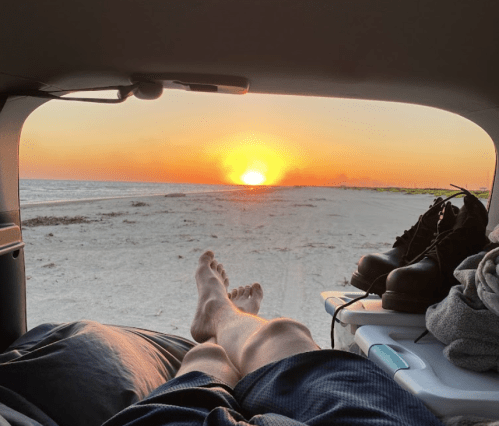 A person relaxes in a car, watching the sunset over a beach, with feet propped up and boots nearby.