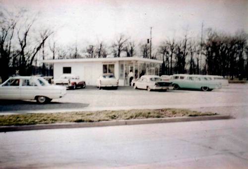 A vintage gas station with several classic cars parked outside, surrounded by bare trees in the background.