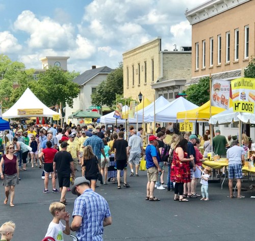 A bustling street market with numerous food stalls and a large crowd enjoying the sunny day.
