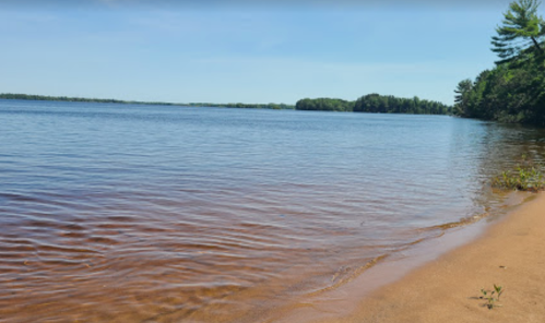 A serene lakeside view with calm water, sandy shore, and lush green trees under a clear blue sky.