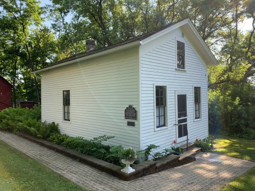 A small white house with a sloped roof, surrounded by greenery and a stone pathway, featuring a historical plaque.