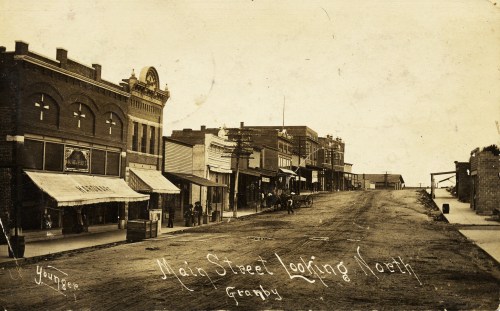 Historic black-and-white photo of Main Street in Granby, showing buildings and a dirt road, looking north.