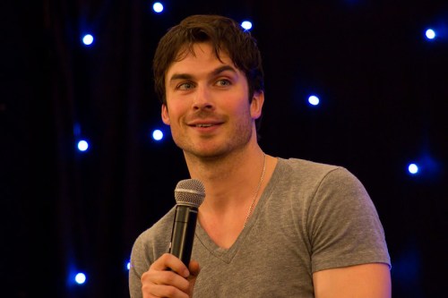 A man with dark hair speaks into a microphone, smiling, against a backdrop of blue lights.