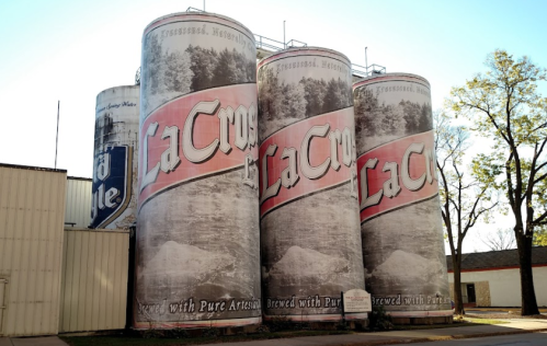 Four large silos wrapped in La Crosse beer advertisements, set against a clear blue sky and trees.
