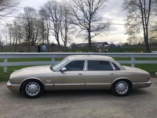 A beige luxury sedan parked on a gravel road, with trees and a farm in the background.