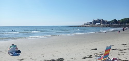 A sunny beach scene with people swimming, a colorful chair, and a building in the background.