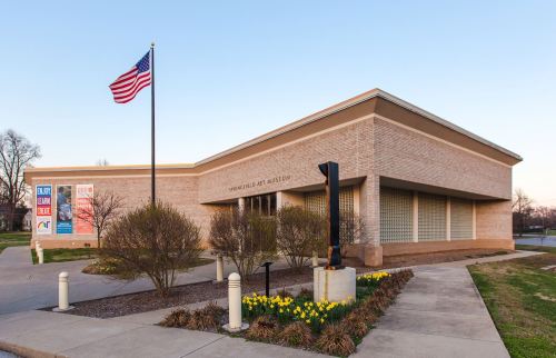 A modern building with a flag, featuring large windows and flower beds in front, set against a clear sky.