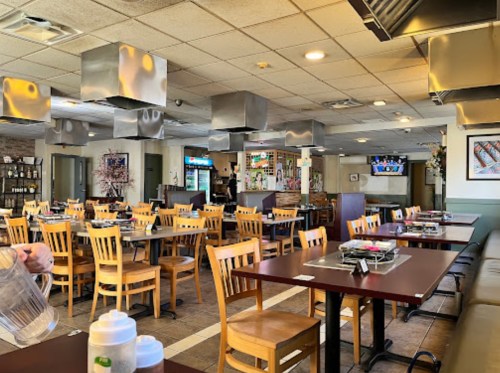 Interior of a spacious restaurant with wooden tables, chairs, and metal ventilation hoods, featuring a bar area in the background.