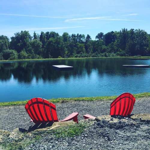 Two red chairs sit by a calm pond, surrounded by greenery and a clear blue sky.