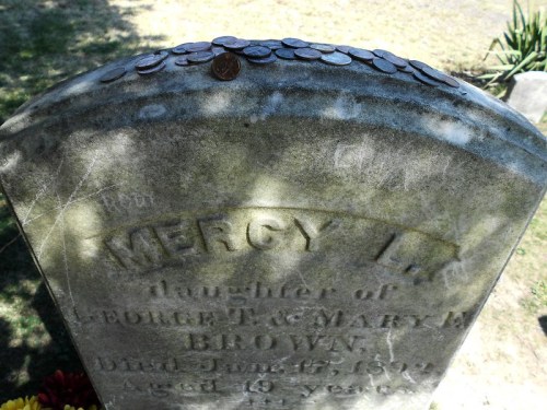 A weathered gravestone with the name "Mercy L." and coins placed on top, surrounded by grass and flowers.