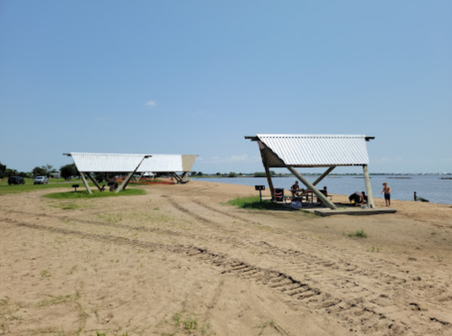 Two modern shelters on a sandy beach by a river, with people enjoying the area under a clear blue sky.