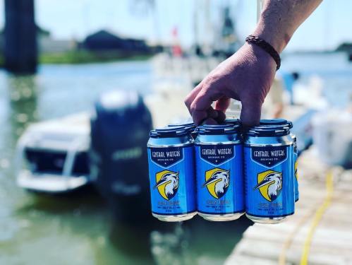 A hand holding a six-pack of Central Waters beer cans by a dock with boats in the background.