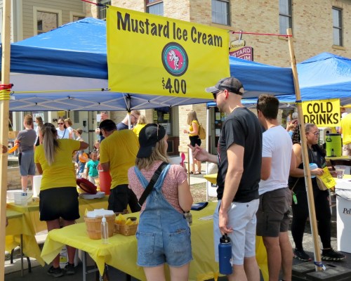 A vibrant outdoor stand selling mustard ice cream, with people in yellow shirts and a sign displaying prices.