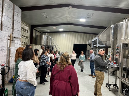 A group of people gathered in a large industrial space, observing a presentation near stainless steel equipment.