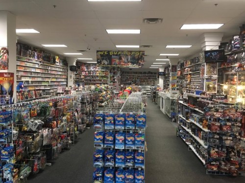 A spacious toy store filled with shelves of action figures, collectibles, and various merchandise. Brightly lit and organized.
