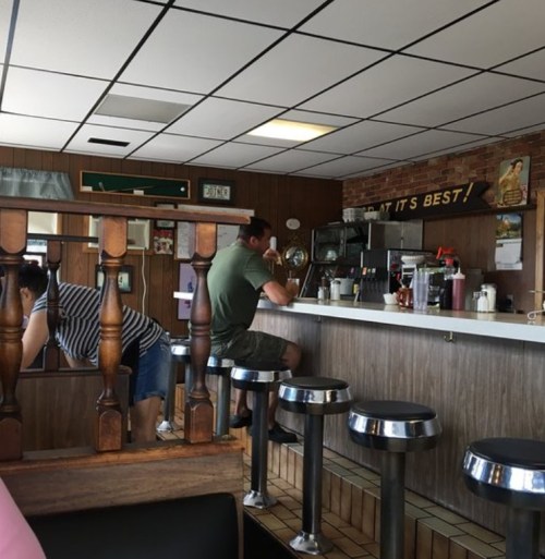 Two people sit at a diner counter, one enjoying a drink while the other leans against the wall in a casual setting.
