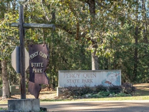 Signage for Percy Quin State Park, featuring a wooden post and stone sign amidst trees and greenery.
