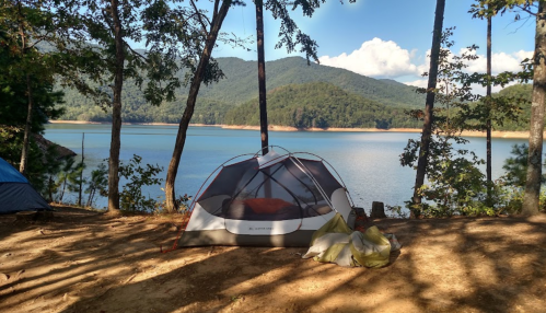A tent set up by a serene lake, surrounded by trees and mountains under a clear blue sky.