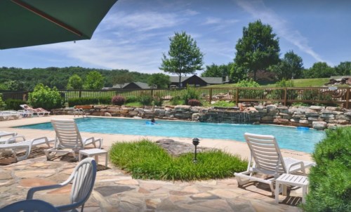 A serene outdoor pool area surrounded by greenery, lounge chairs, and a stone waterfall under a clear blue sky.